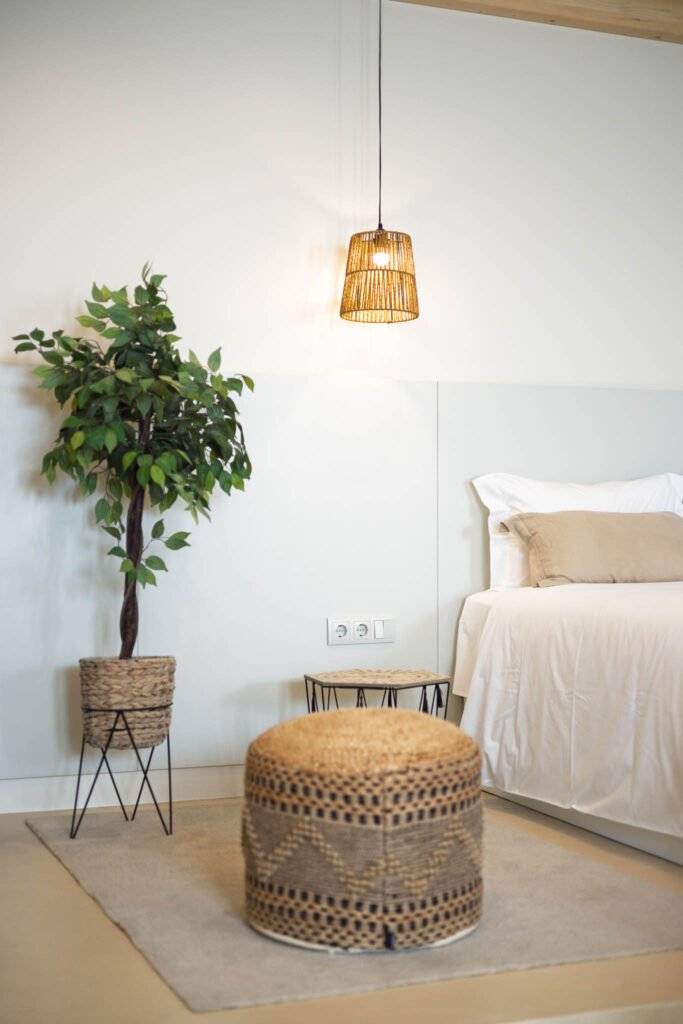 Detalhe de pufe em fibra natural, planta em vaso e candeeiro suspenso junto à cama em alojamento local