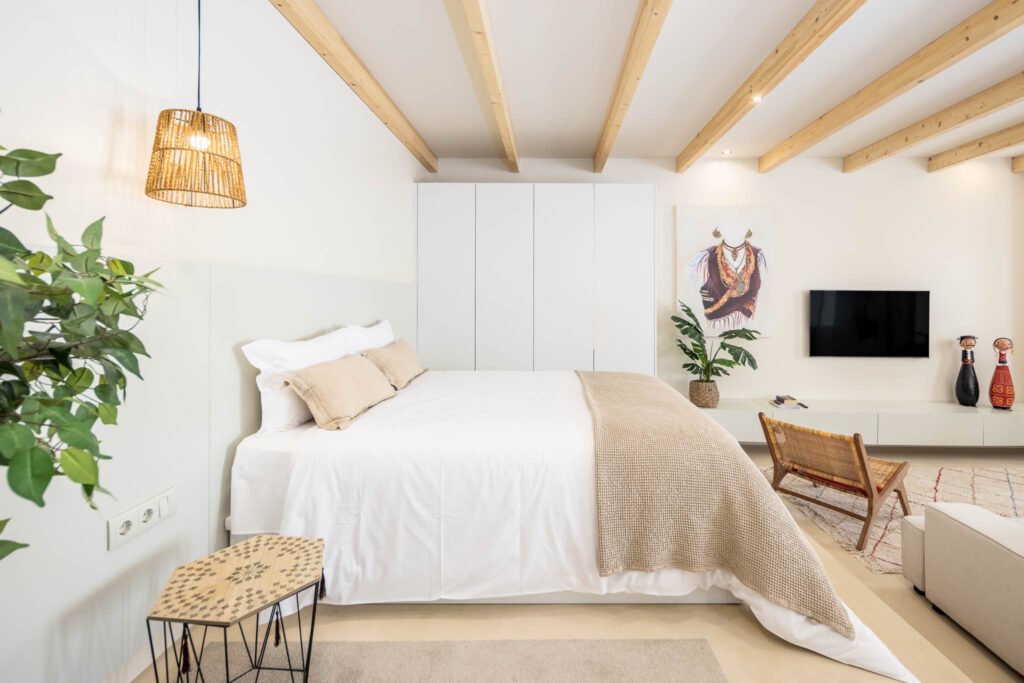 Cama de casal sob vigas de madeira à vista, candeeiro em fibra natural, roupeiro branco e tela de traje tradicional