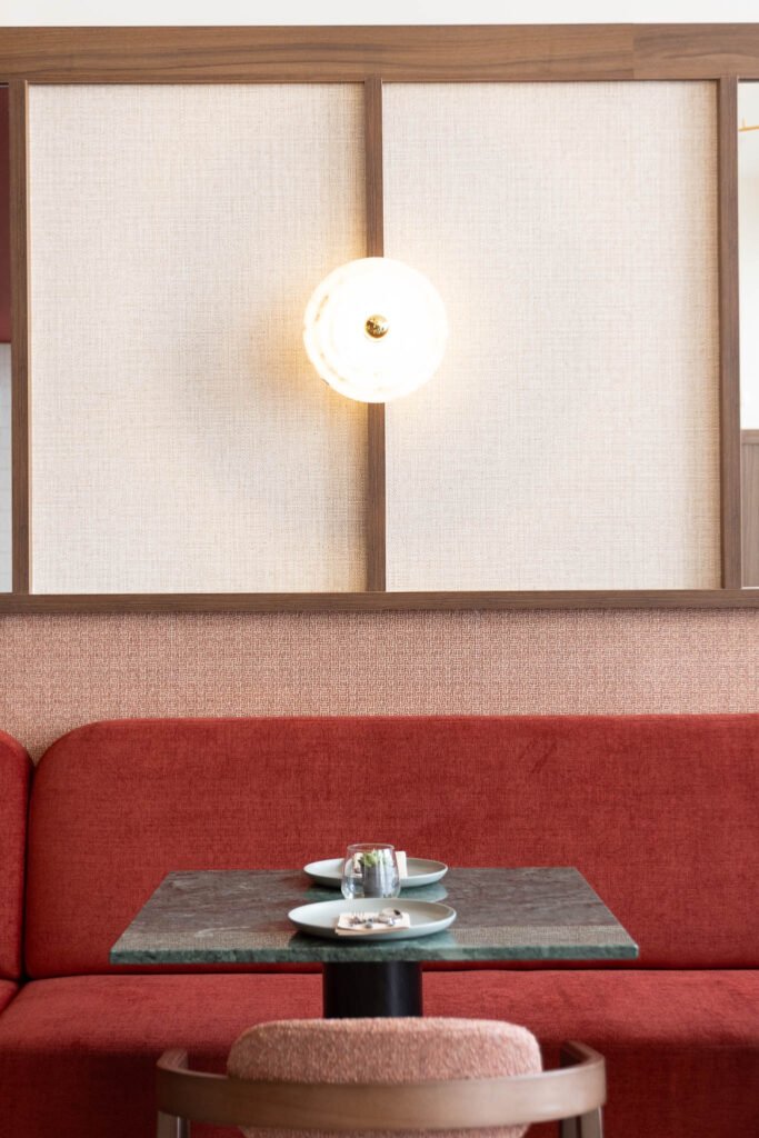 Detalhe de interior de hotel boutique: mesa de tampo verde junto a banco corrido vermelho, sob aplique de luz entre painéis emoldurados em madeira
