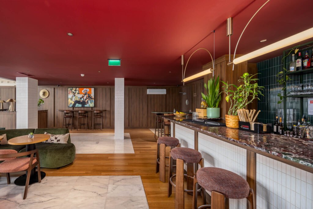 Bar de hotel boutique com tecto vermelho, pendentes lineares, bancos estofados e zona de estar com sofá verde ao fundo