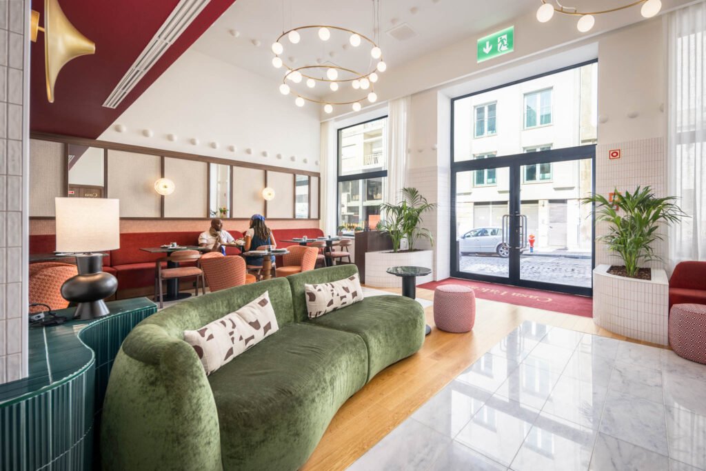 Zona de pequeno-almoço e lobby da Casa Falcão, com sofá verde em primeiro plano e porta envidraçada aberta para a rua